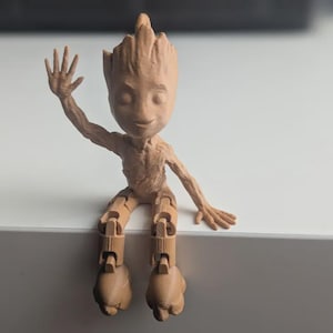 May include: A tan-colored articulated figure of a character with a tree-like appearance, seated with one arm raised in a wave. The figure has a detailed texture and is posed on a white surface.