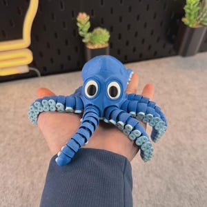May include: A blue octopus figurine with large white eyes and light blue accents on its tentacles. The octopus is held in a hand, showcasing its detailed design. The figurine is a whimsical decorative item.