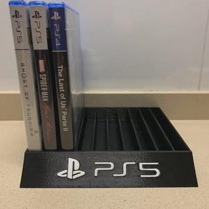 May include: Black PS5 game holder with white PS5 logo. Three game cases are displayed: "Ghost of Tsushima", "Marvel Spider-Man Miles Morales", and "The Last of Us Parte II". The PS4 game case is also visible.