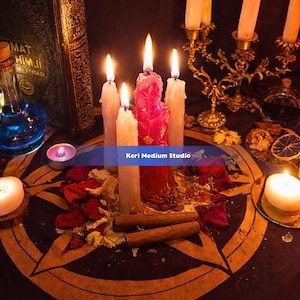 May include: A mystical scene featuring lit candles of various sizes and colors, including a central red candle. The arrangement includes a pentagram, dried flowers, cinnamon sticks, and a vintage book. The text "Keri Medium Studio" is visible.