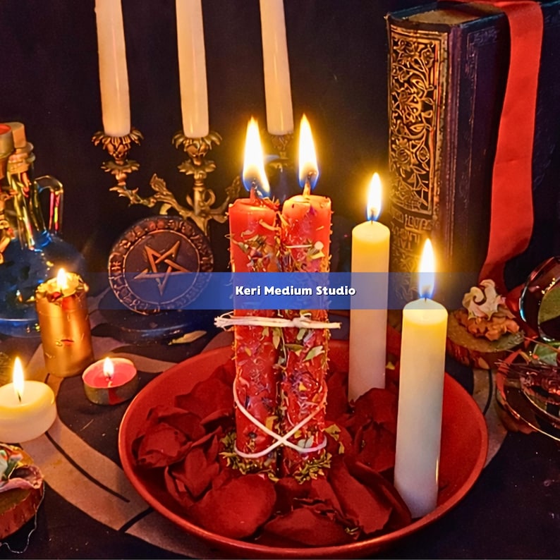 May include: A collection of lit candles, including two red candles adorned with herbs and tied together, and several white candles. The candles are arranged with a red bowl of rose petals, a decorative brass candelabra, and a book. The text "Keri Medium Studio" is visible.