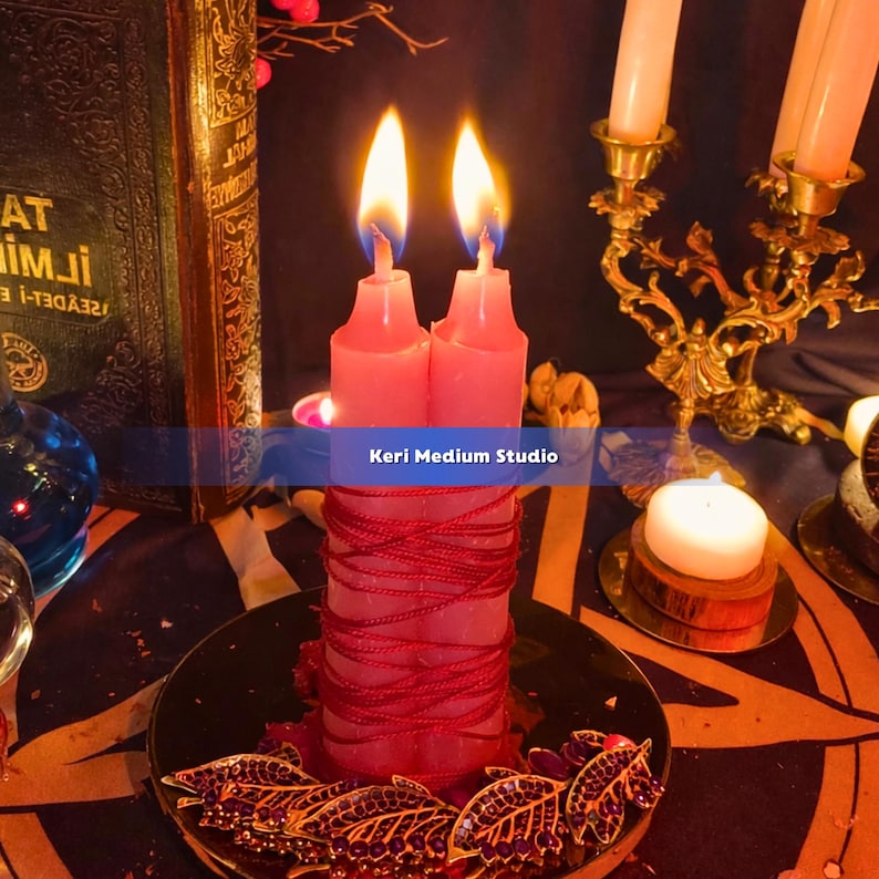 May include: A pair of pink candles with flames, wrapped with red string, sit on a decorative plate. The plate has a gold leaf design. The background includes a vintage book, a gold candle holder, and the text "Keri Medium Studio".