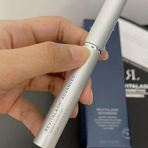 Könnte beinhalten: Ein silbernes Wimpernconditioner-Tübchen wird gehalten. Auf der Tube steht "REVITALASH® ADVANCED" und "eyelash conditioner 3,5 ml / 0,118 fl oz". Im Hintergrund ist eine dunkelblaue Schachtel zu sehen.