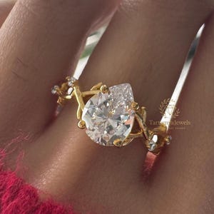 May include: A gold ring featuring a pear-shaped, clear gemstone. The ring has a delicate, floral design with small accent stones. The ring is shown on a hand wearing a red sweater. The brand name "Tara Jewels" is visible.