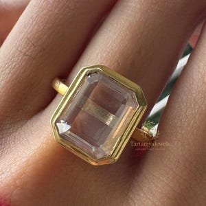Könnte beinhalten: Ein goldener Ring mit einem großen, rechteckigen, klaren Edelstein und einem goldenen Band. Der Ring hat eine goldene Lünettenfassung. Der Ring befindet sich am Finger einer Person.