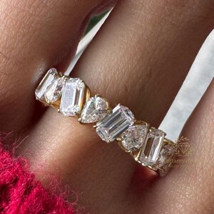Könnte beinhalten: Ein goldener Ring mit einer Reihe klarer, rechteckiger und herzförmiger Steine. Der Ring wird an einem Finger getragen, mit einem leuchtend rosa Pullover im Hintergrund. Der Ring ist ein Schmuckstück.