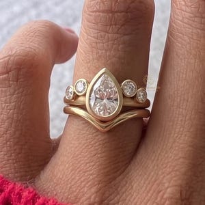 Könnte beinhalten: Ein goldener Ringsatz mit einem birnenförmigen Diamanten als Mittelstein und einem geschwungenen Band mit kleineren runden Diamanten. Der Ring wird mit einem passenden Band mit zusätzlichen runden Diamanten kombiniert.