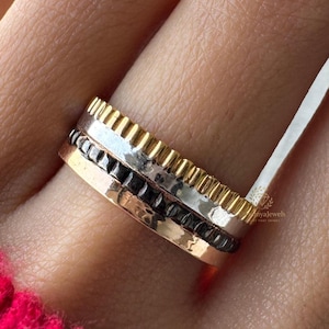 Könnte beinhalten: Nahaufnahme von drei gestapelten Ringen an einem Finger. Die Ringe sind goldfarben, silberfarben und schwarz. Die oberen und unteren Ringe haben ein strukturiertes, geriffeltes Design. Der mittlere Ring ist schwarz mit einer gehämmerten Textur. Die Person trägt einen leuchtend rosa Pullover.