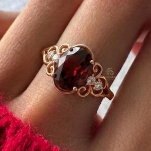 Könnte beinhalten: Roségoldener Ring mit einem großen, oval geschliffenen, tiefroten Edelstein. Der Ring hat ein dekoratives Filigran-Design mit kleinen, klaren Akzentsteinen. Der Ring ist vor rotem Hintergrund platziert.