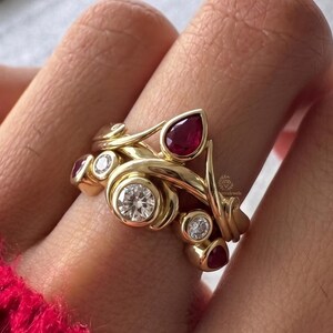 Peut inclure: Une bague en or au design unique et organique. Elle présente une grosse pierre précieuse claire et ronde, plusieurs petites pierres précieuses claires et des pierres précieuses rouges en forme de goutte. La bague est portée sur un doigt, avec un pull rouge en arrière-plan.