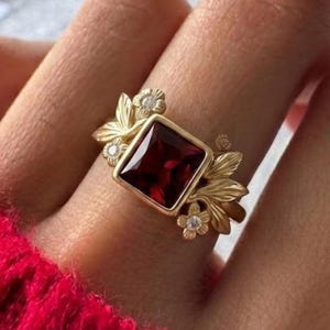 Könnte beinhalten: Ein goldener Ring mit einem quadratischen, tiefroten Edelstein. Der Ring ist mit Blatt- und Blumendetails sowie kleinen, klaren Akzentsteinen verziert. Der Ring wird an einem Finger gezeigt, mit einem roten Pullover im Hintergrund.