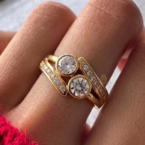 Könnte beinhalten: Ein goldener Ring mit zwei markanten, rund geschliffenen, klaren Steinen in einem Bypass-Design. Das Band ist entlang der geteilten Ringschiene mit kleineren, im Pavé-Stil gefassten klaren Steinen besetzt, was eine geschichtete Optik mit poliertem Metallfinish erzeugt.