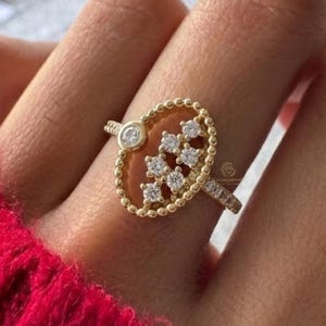 Könnte beinhalten: Ein goldener Ring mit einem ovalen Design, das mit kleinen, klaren Steinen besetzt ist. Der Ring hat einen Perlenrand und kleine Steine am Band. Der Ring wird auf einer Hand vor dem Hintergrund eines roten Pullovers gezeigt.