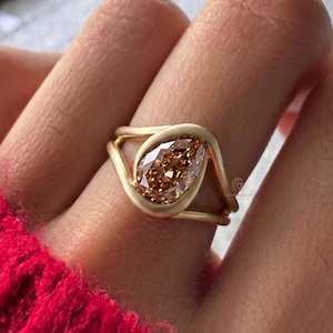 Könnte beinhalten: Ein goldener Ring mit einem birnenförmigen, hellbraunen Edelstein. Das Band des Rings hat ein einzigartiges, offenes Design, das sich um den Edelstein legt. Der Ring ist vor dem Hintergrund einer Hand und eines roten Pullovers abgebildet.