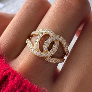 Könnte beinhalten: Ein goldfarbener Ring mit einem Doppel-C-Design, besetzt mit kleinen, klaren Edelsteinen. Der Ring wird vor einem roten Pullover gehalten, wodurch das Funkeln und Design des Schmucks hervorgehoben werden. Der Ring ist ein Modeaccessoire.