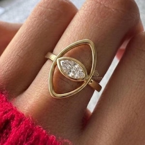 Könnte beinhalten: Ein goldener Ring mit einem Diamanten im Marquiseschliff, der in einem ovalen Rahmen gefasst ist. Der Ring wird an einem Finger getragen und unterstreicht sein elegantes Design und seinen Glanz. Der Ring hat eine polierte Oberfläche.