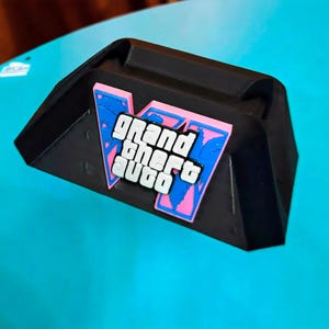 May include: A black plastic stand featuring a prominent Grand Theft Auto V logo emblem. The emblem displays the white text Grand Theft Auto over a stylized blue and pink V shape. The stand has a wide, angled base and a recessed top section.
