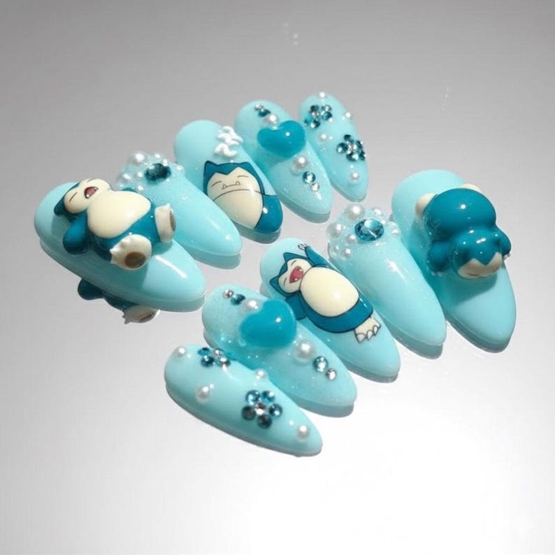 May include: Light blue acrylic nails with various designs. Some nails feature a 3D Snorlax figure, while others have heart-shaped accents, rhinestones, and pearl embellishments. The nails are oval-shaped and have a glossy finish.