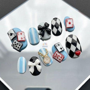 May include: A set of ten artificial nails with an Alice in Wonderland theme. Designs include playing cards, black and white checkered patterns, bows, and a pocket watch. The nails are painted in shades of blue, white, and black.