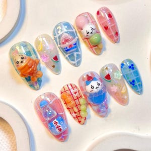 May include: A set of ten almond-shaped, decorated fingernails. Each nail features a different design, including cartoon characters, fruit, and Japanese text. Colors include pink, blue, orange, and green. The nails have a glossy finish and are set against a white background.