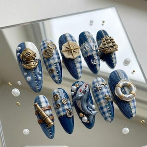 May include: A set of ten press-on nails featuring a nautical theme with blue and white gingham patterns and denim textures. Embellishments include a gold diving helmet, gears, a compass, anchors, a ship, a telescope, buttons, a shark, and a lifebuoy.