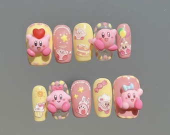 May include: A set of ten artificial nails with a cartoon theme. Each nail features a different design, including a pink character with a heart, stars, and a cupcake. The nails are painted in shades of pink and yellow.