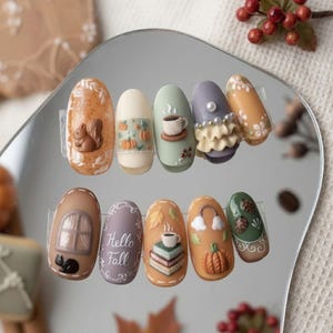 May include: A set of ten press-on nails featuring autumn-themed 3D designs including a squirrel, pumpkins, a coffee cup, pearls, a window with a black cat, the text Hello Fall, a stack of books, and pinecones in shades of orange, green, and purple.