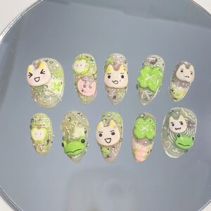 May include: A collection of ten false nails, each decorated with cartoon characters and designs. The nails feature smiling cat faces, green frogs, and four-leaf clovers in 3D. The colour palette includes green, pink, and white. The nails are displayed on a reflective surface.