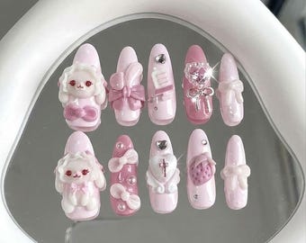 May include: A collection of eleven artificial nails in shades of pink. Some nails are decorated with 3D bows, hearts, and a bunny. Others feature rhinestones or a strawberry design. The nails are displayed on a reflective surface.