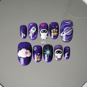 May include: A set of ten press-on nails with a dark purple glitter base. Each nail features a different space-themed design, including a rocket, a planet, a cloud, and cartoon characters. The designs are in white, pink, and blue.