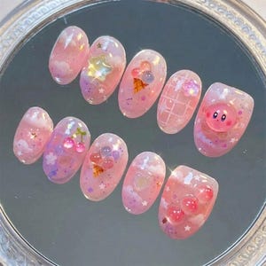 May include: A set of ten pink and clear acrylic nails with various designs. The designs include clouds, stars, cherries, ice cream cones, and a cartoon character. The nails are oval-shaped and have a glossy finish.