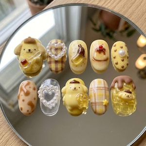 May include: A collection of ten artificial nails with various designs. The nails feature yellow, brown, and clear bases with detailed decorations, including cartoon characters, fruit, plaid patterns, and pearl accents. The nails are arranged on a reflective surface.