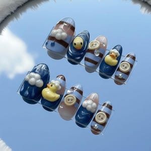May include: A set of ten artificial nails with various designs. Some nails feature a blue base with white cloud accents, while others have a striped pattern. Several nails showcase a yellow rubber ducky or a button.
