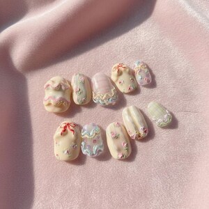 May include: A collection of ten artificial nails in various designs. The nails feature pastel colors like pink, cream, and light blue, with decorative elements such as floral patterns, bows, and wavy lines. The nails are arranged on a pink fabric surface.
