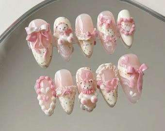 Puede incluir: Un conjunto de diez uñas artificiales en forma de almendra. Las uñas presentan una base rosa claro con elementos decorativos blancos y rosas, incluyendo lazos, ositos de peluche y encaje. Algunas uñas tienen un diseño moteado, mientras que otras son de color rosa liso.