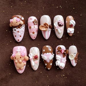 May include: Ten almond-shaped artificial nails, each with a unique design. The nails feature pink, brown, and white colours, decorated with 3D embellishments such as hearts, bows, and cartoon characters. Some nails have musical notes and stars.