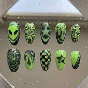 May include: A set of ten press-on nails featuring a vibrant green and black color palette. Designs include an alien face, black spirals, stars, dripping slime patterns, a black and green checkerboard, and smiley faces, all accented with glitter.