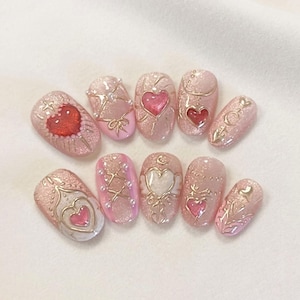 Puede incluir: Un conjunto de diez u&ntilde;as artificiales con una base de purpurina rosa. Cada u&ntilde;a presenta un dise&ntilde;o diferente, incluyendo corazones, lazos y adornos dorados. Algunas u&ntilde;as tienen un dise&ntilde;o de coraz&oacute;n rojo, otras un coraz&oacute;n rosa.
