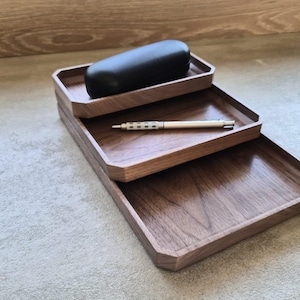 May include: Three rectangular wooden trays with beveled edges stacked in a tiered arrangement. A black oval case rests on the top tray, and a silver metallic pen with a textured grip lies horizontally across the middle tray.