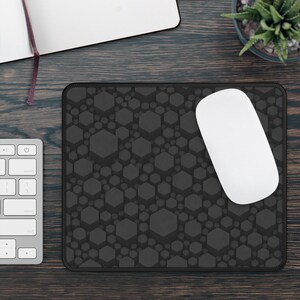 May include: A rectangular mouse pad featuring a dark grey and black geometric pattern of various sized hexagons. The mouse pad has a stitched black border and a smooth surface texture, providing a consistent base for a white computer mouse.