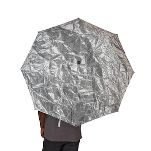 May include: A silver umbrella with a highly reflective, crinkled metallic texture. The canopy is octagonal in shape and features a small black circular detail at the centre. The umbrella is held open, displaying a distinct, uneven surface pattern.