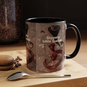 Könnte beinhalten: Eine Keramiktasse mit schwarzem Innenraum und Henkel, verziert mit einem sich wiederholenden Muster von Schlangen in verschiedenen Farben. Auf der Tasse steht der Text "Slippery Little Sucker". Die Tasse steht auf einer Holzoberfläche mit einem Löffel und einem Gebäck.