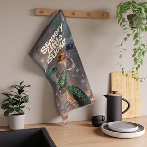 Op de afbeelding: Een holografische keukendoek met de tekst "Slippery Little Sucker" en illustraties van drie slangen in groen, goud en blauw. De doek hangt aan een houten rek.