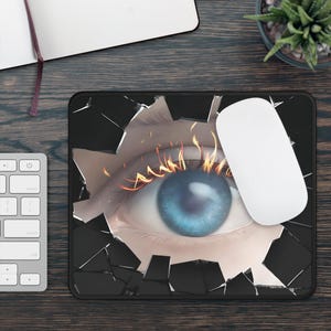 May include: Black rectangular mousepad featuring a blue eye with flames around the upper eyelid, breaking through a shattered glass effect. A white wireless mouse rests on the right side of the pad. The design is set against a dark background.
