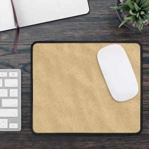 May include: A rectangular mouse pad with a textured, light tan, sand-coloured surface and a solid black stitched border. The mouse pad is positioned on a dark wood grain surface next to a white computer mouse and a white keyboard.