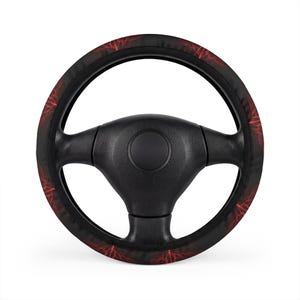 Peut inclure: Un couvre-volant noir orné d'un motif répétitif d'éclairs rouges. La housse est installée sur un volant noir standard à trois branches, doté d'un bouton de klaxon circulaire central et de surfaces de préhension texturées sur les branches.