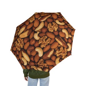 May include: A collapsible umbrella featuring a repeating pattern of various nuts, including brown almonds, tan cashews, and textured walnuts. The canopy displays a dense, realistic photographic print of mixed nuts covering the entire surface area.
