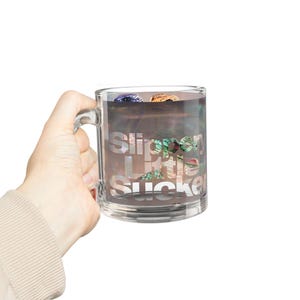 Puede incluir: Una taza de vidrio transparente con las palabras "Slippery Little Sucker" en una fuente blanca y estilizada. La taza contiene un diseño oscuro e iridiscente con toques de azul y naranja.