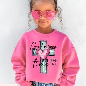 May include: A pink sweatshirt with the text "God is Good All the Time" and a cross design with a heart in the center. The sweatshirt is labeled "Safety Pink" and is worn by a young person wearing pink heart-shaped sunglasses.