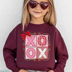 Peut inclure: Un sweat-shirt bordeaux avec un motif décoratif "XO". Le motif présente des motifs roses et rouges, notamment des cœurs, du vichy et des rayures. Le sweat-shirt porte les inscriptions "Tiny Baby Love" et "MAROON".